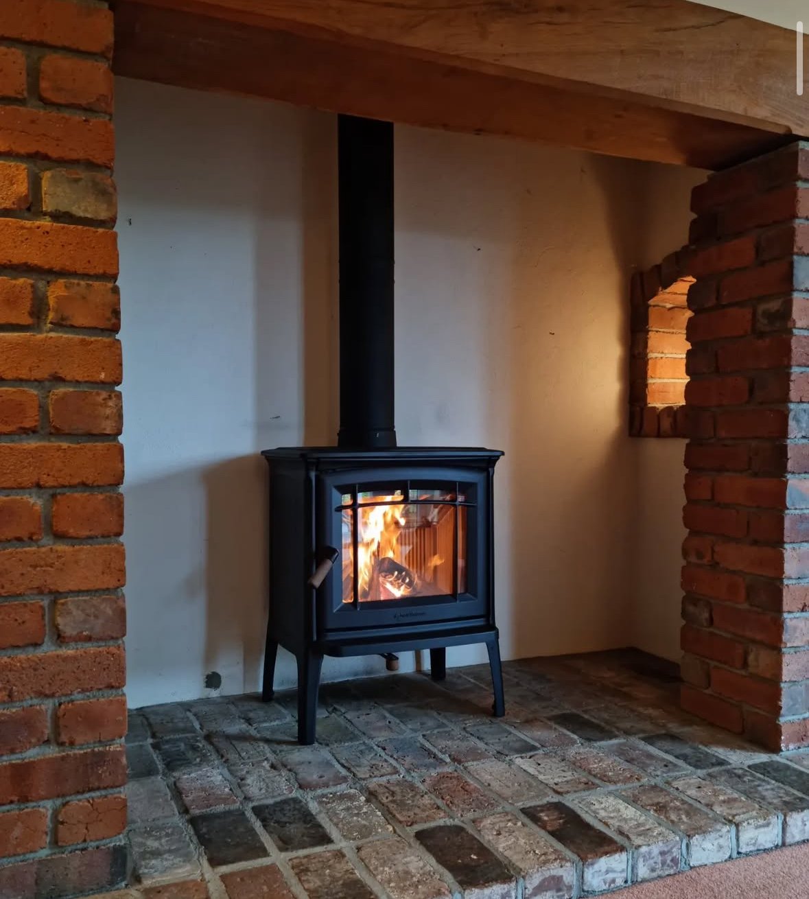 A black wood-burning stove with a glass door showing a burning fire inside, set on a brick hearth with a brick wall on the left and a small brick window opening on the right, inside a cozy room.