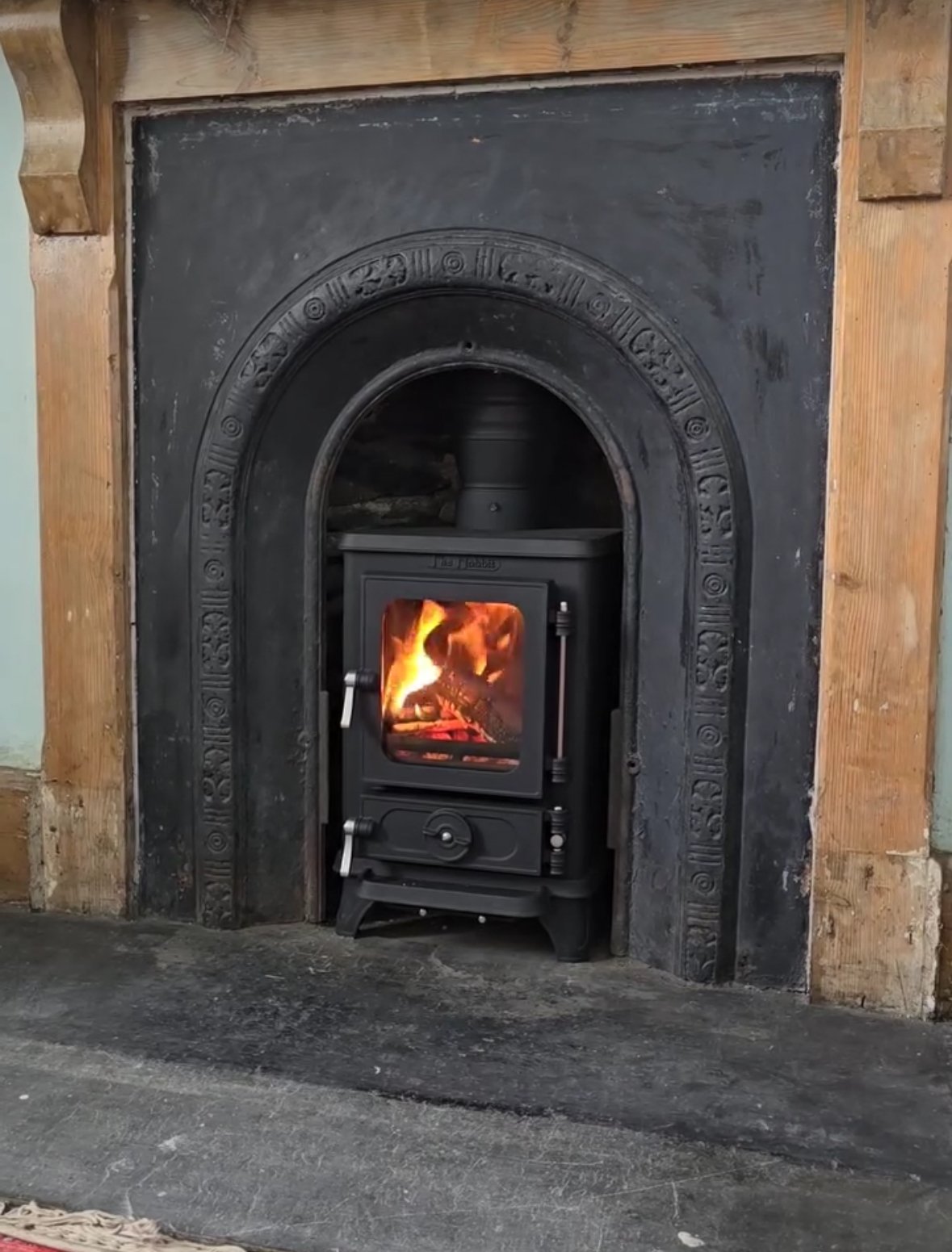 A wood-burning stove with fire inside, set in a black ornate fireplace with wooden trim around it.