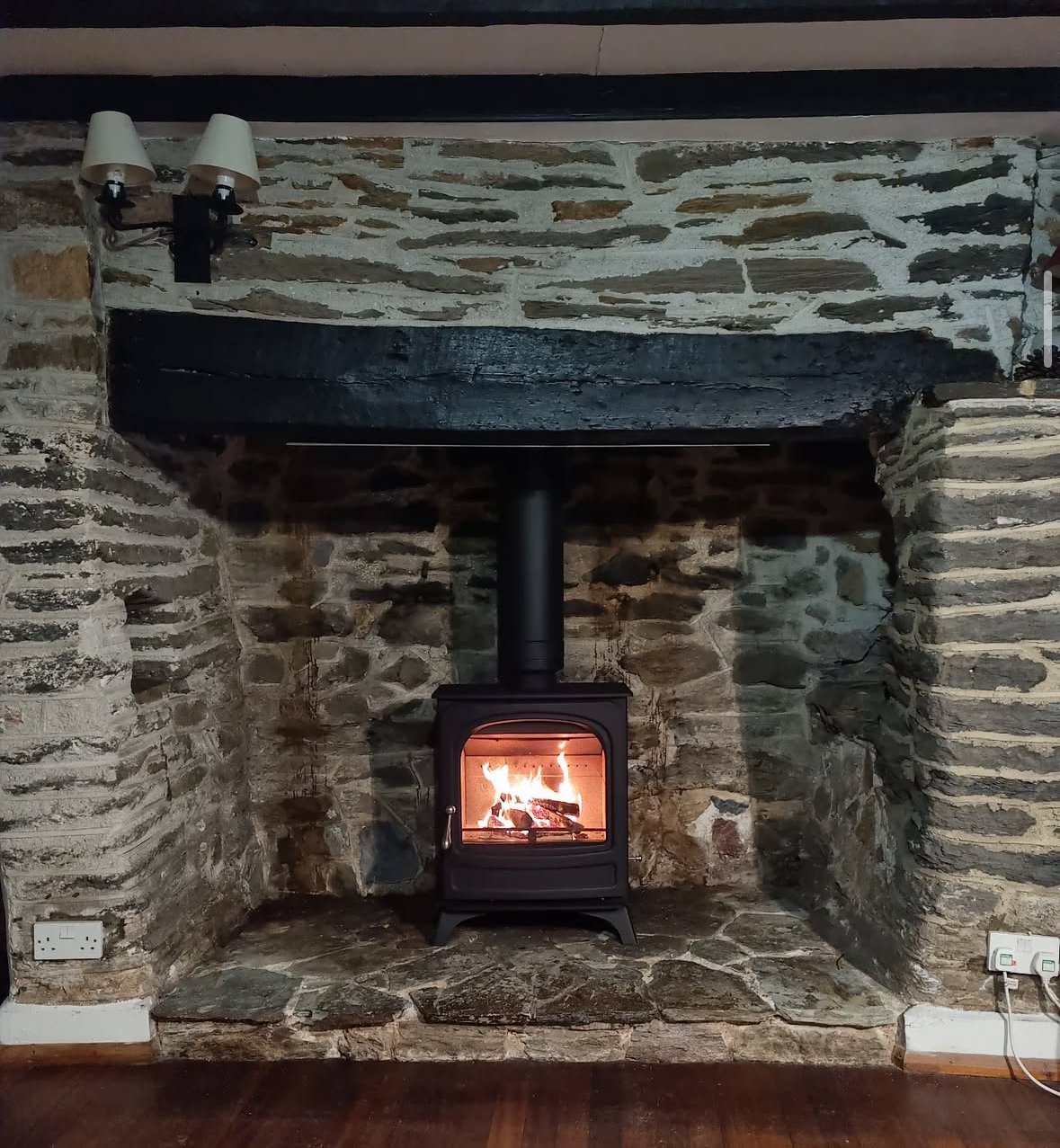 Stone fireplace with a black wood stove burning inside, set in a rustic room with exposed stone walls and a wooden mantel.