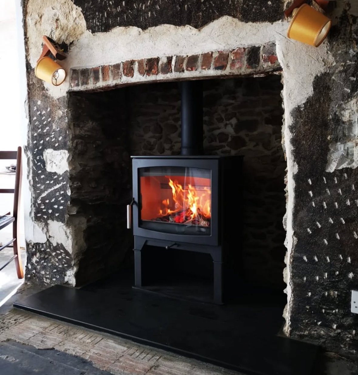 Wood stove with a fire burning inside, set in a rustic stone and brick fireplace surrounded by rough, unfinished black and white plaster walls.