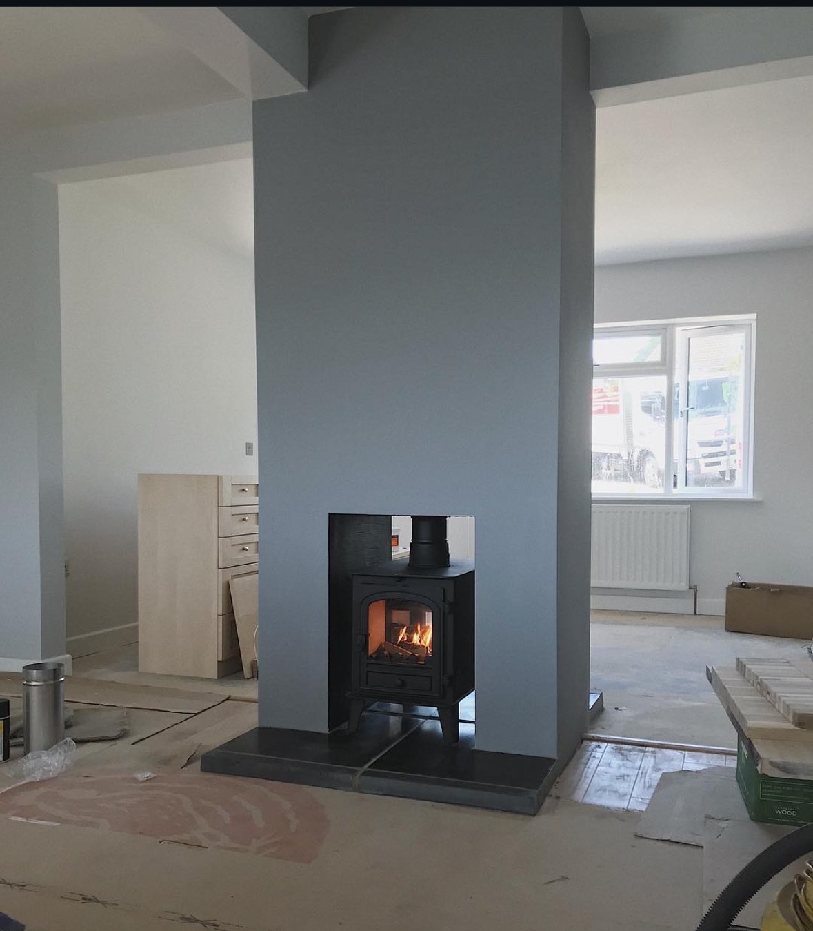 Interior of a house under construction with a black wood stove burning on a black tile hearth, a light blue wall behind it, a window with cars outside, and various construction materials on the floor.