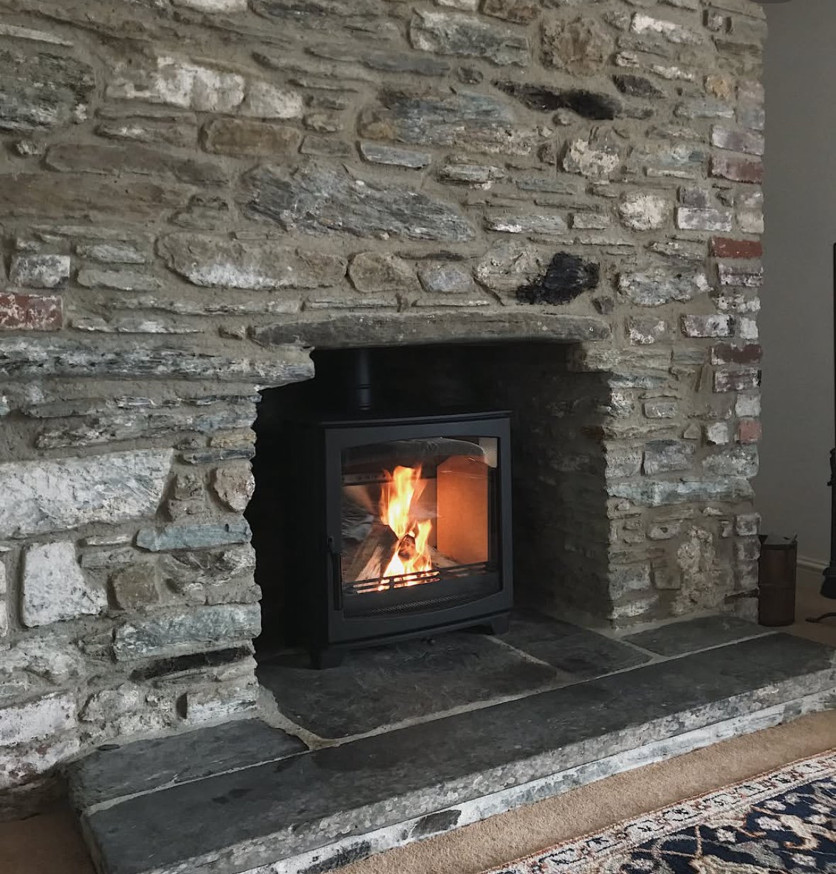 A cozy stone fireplace with a fire burning inside. The fireplace is made of rough, uneven stones and has a black metal insert. The hearth is also tiled with large irregular stones, and a patterned rug is partially visible in the foreground.