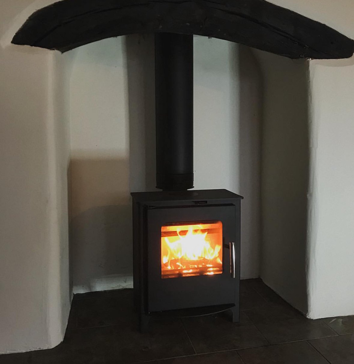 Black wood stove with glass door burning with flames, set against a textured white wall with a black chimney pipe extending upward.