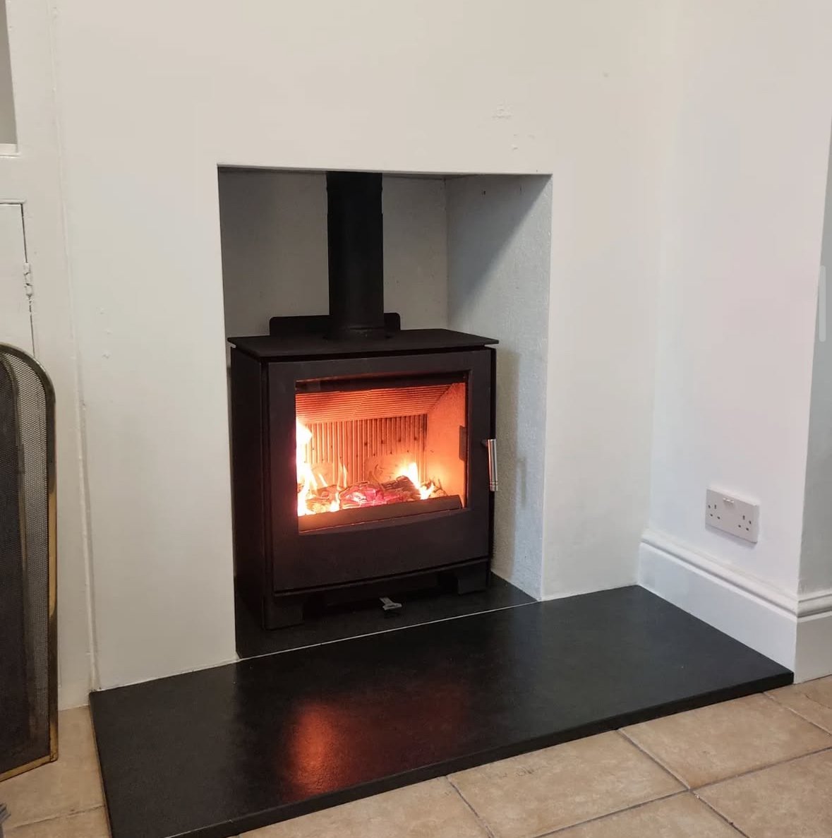 A black wood stove with a glass front, burning with visible flames and logs inside, is set into a white wall fireplace with a black hearth and black chimney above.