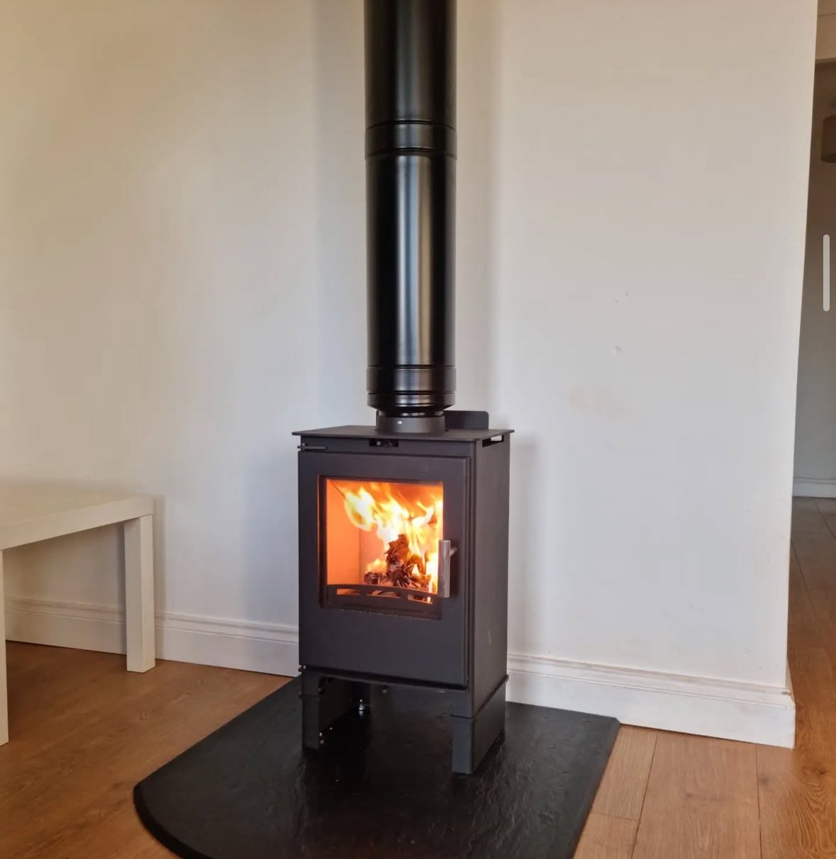 A black wood-burning stove with a glass front showing a fire inside, placed on a black mat against a white wall, with a black chimney pipe extending upward.