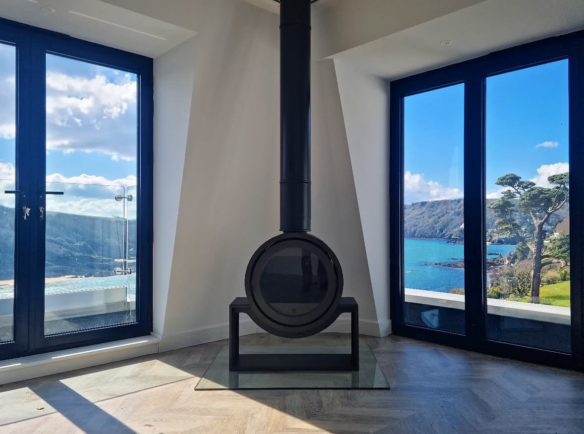 Modern living room with a black hanging fireplace, large windows showing a lakeside view with trees, cliffs, and blue sky.