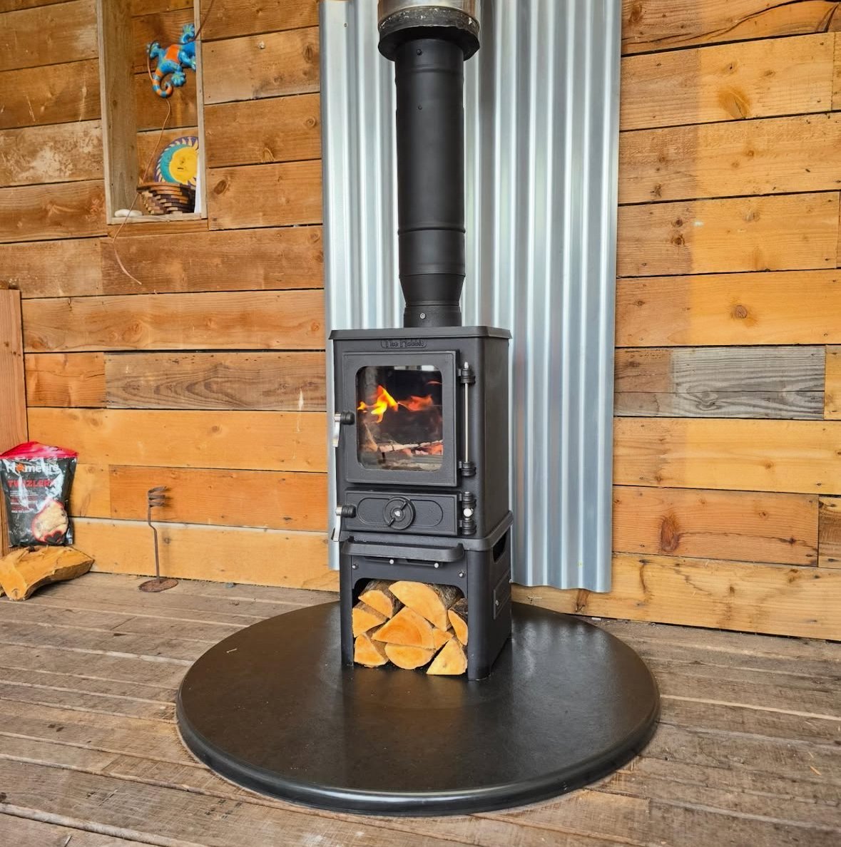 A black wood stove with a visible fire inside, connected to a black chimney pipe, sits on a black circular pad in a wooden room with horizontal wood paneling and a corrugated metal backdrop.