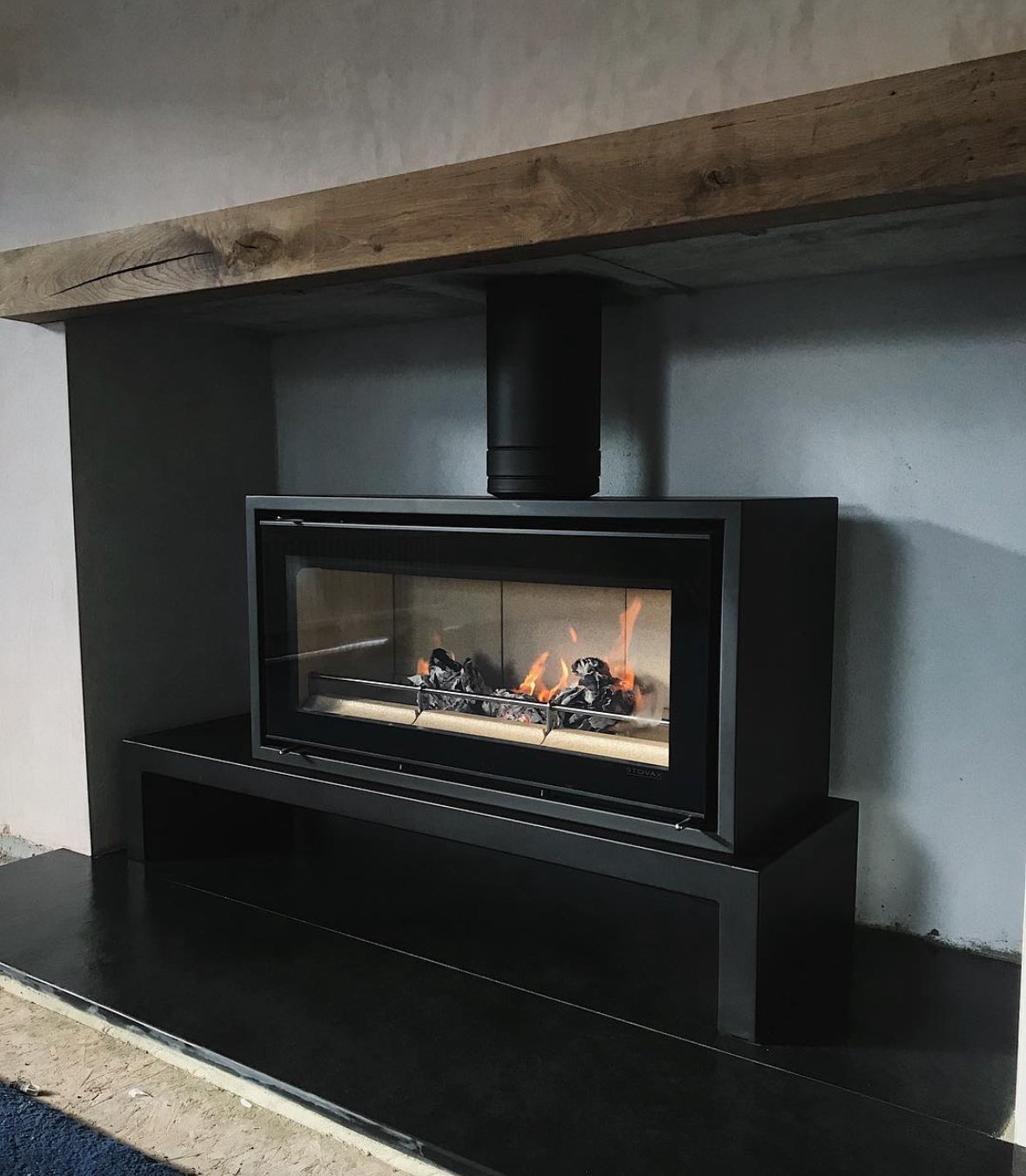 Modern black rectangular fireplace with a glass front, set into a wall with a gray background and a wooden beam above.