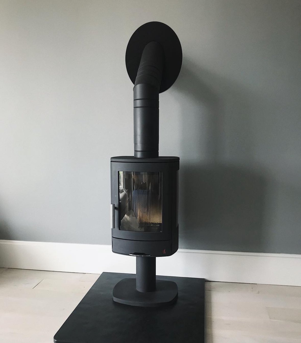 Black wood stove with a glass door, pipe leading to a black wall vent, positioned on a black mat against a light gray wall.
