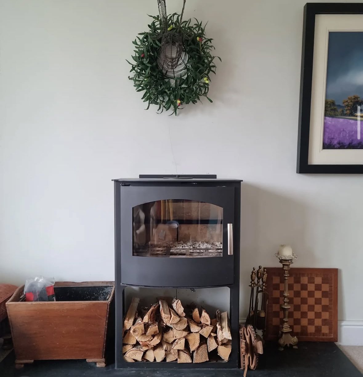 A fireplace with firewood stacked underneath, a decorative candle holder with a candle to the right, and a framed picture of a lavender field on the white wall above, with a wall wreath hanging above the fireplace.