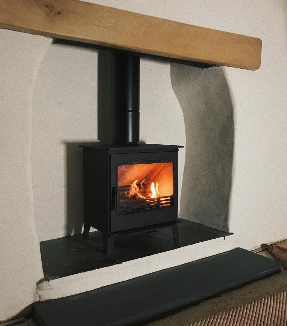 A black wood stove with glass door, burning fire inside, set into a white wall alcove with a dark hearth. A wooden beam extends across the top of the alcove.