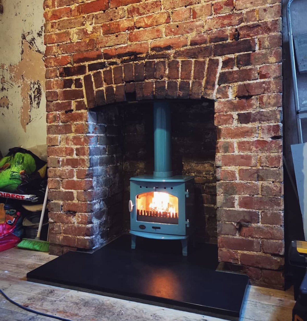 A cozy room featuring a brick fireplace with a small wood-burning stove inside, burning with bright flames. To the left, cleaning supplies and a green bag are visible. To the right, part of a shelving unit can be seen.