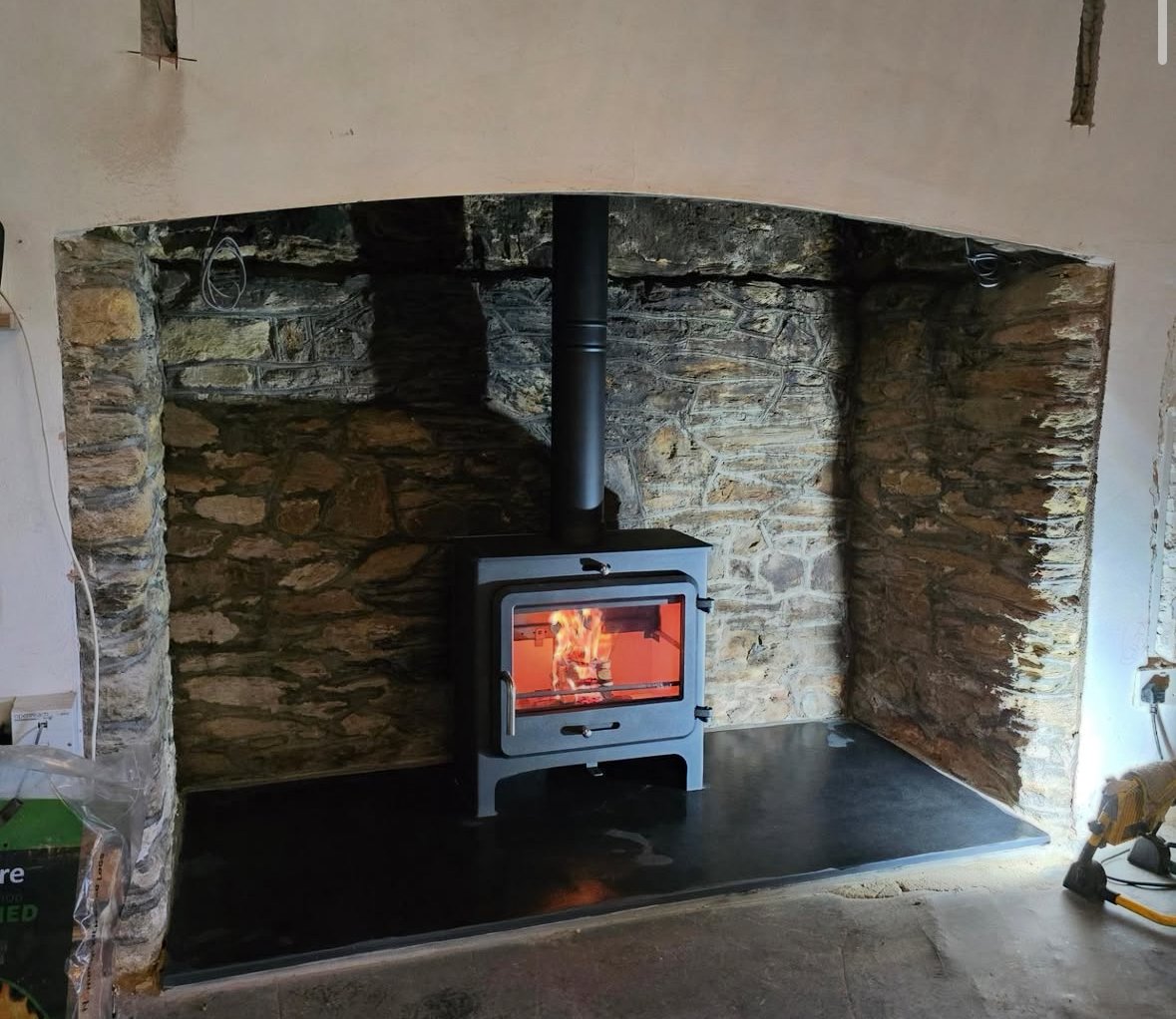 A wood stove with visible flames inside, set against a stone wall in a room under construction or renovation. Construction tools and materials are nearby.