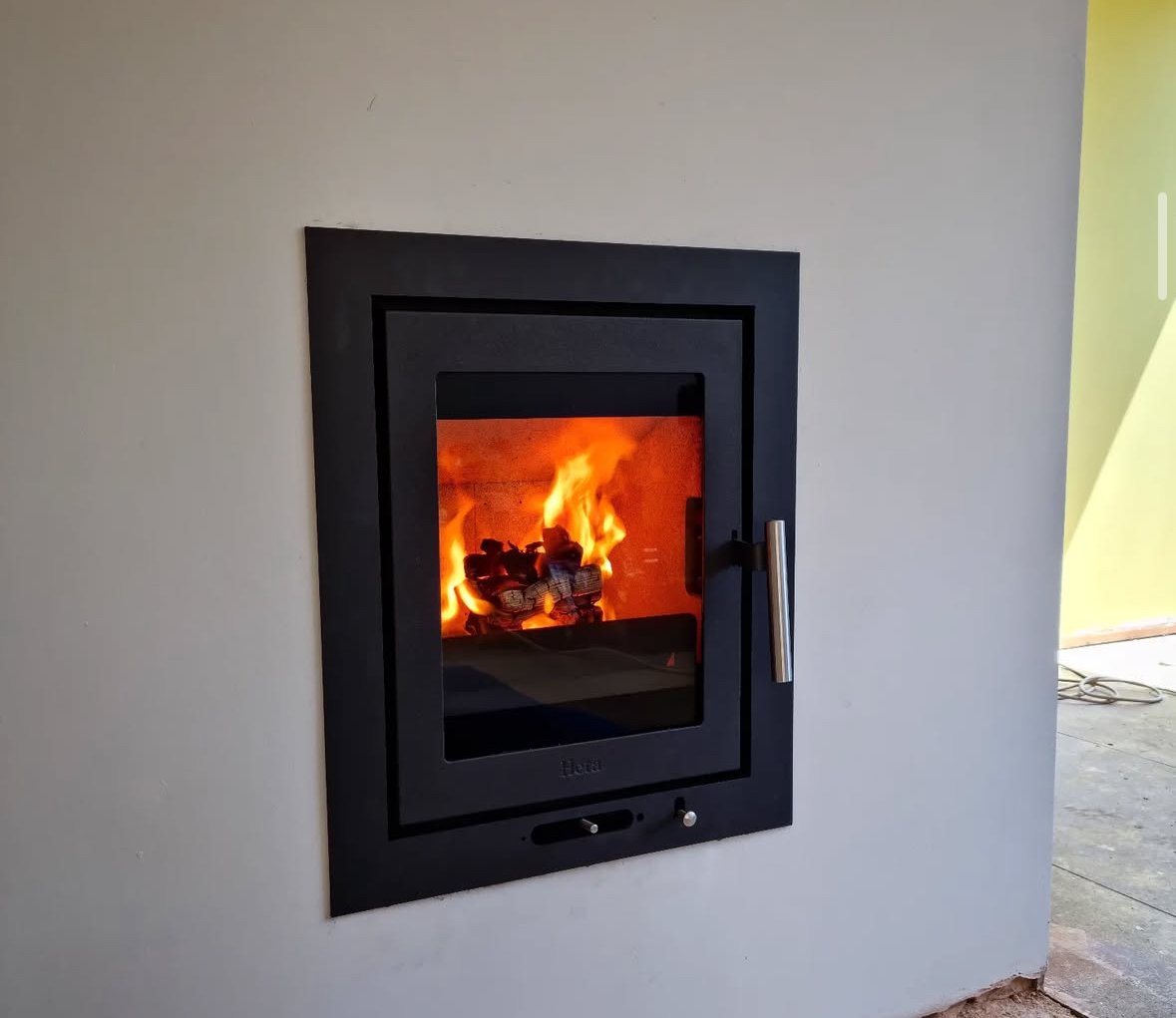 A modern built-in fireplace with a glass door, glowing with orange flames and burning logs inside, set into a white wall.