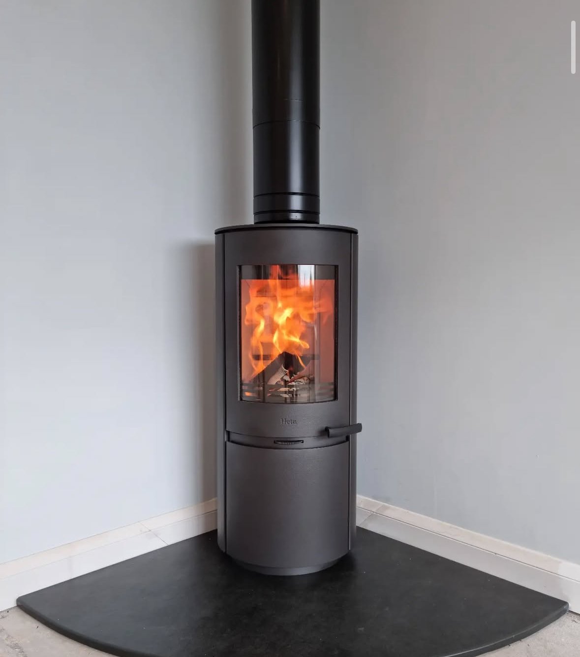 Black wood-burning stove with visible flames inside, connected to a chimney pipe, located on a black hearth in a room with light-colored walls.