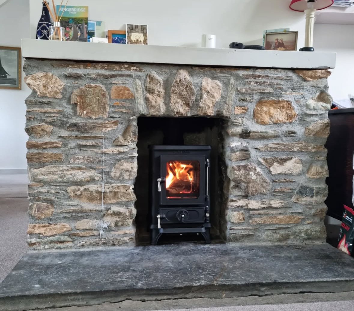 A stone fireplace with a small black stove inside, fire visible through the glass door, and a white mantle on top with various picture frames and decorative items.