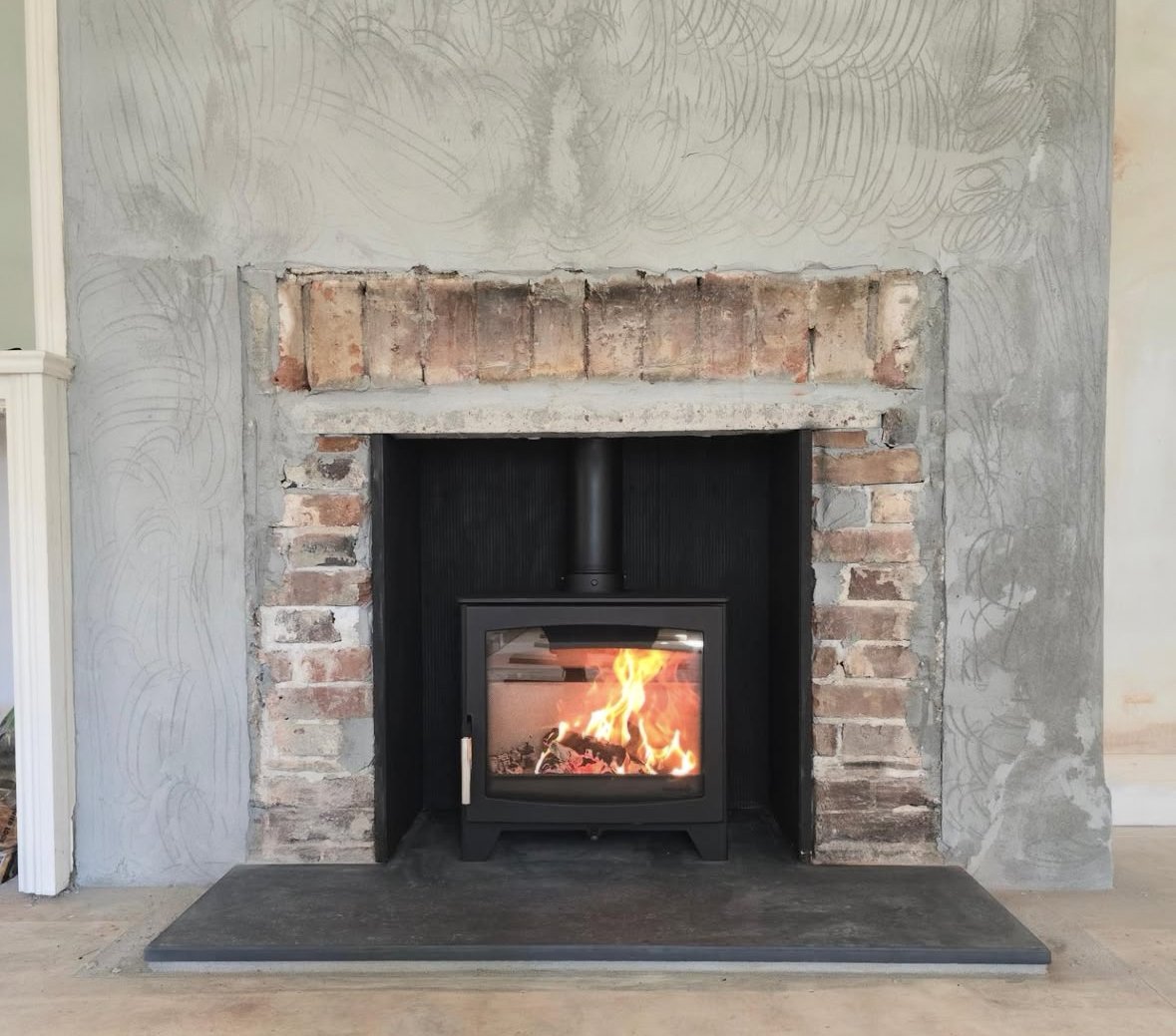 A fireplace with a black stove insert burning with flames inside, surrounded by unfinished brick and concrete wall.