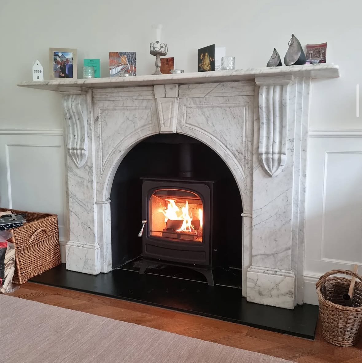White marble fireplace with a lit wood stove, decorated with various small objects and photographs on the mantelpiece, a wicker basket on the floor to the left, and a woven basket to the right.