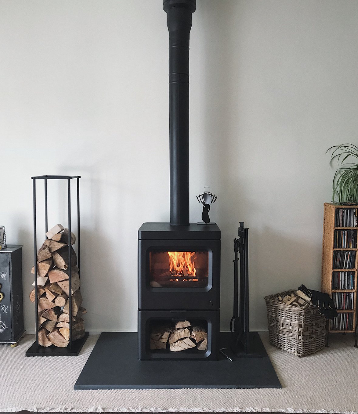 Wood stove with a fire burning, surrounded by firewood storage, fireplace tools, a plant, and shelves with books or CDs.