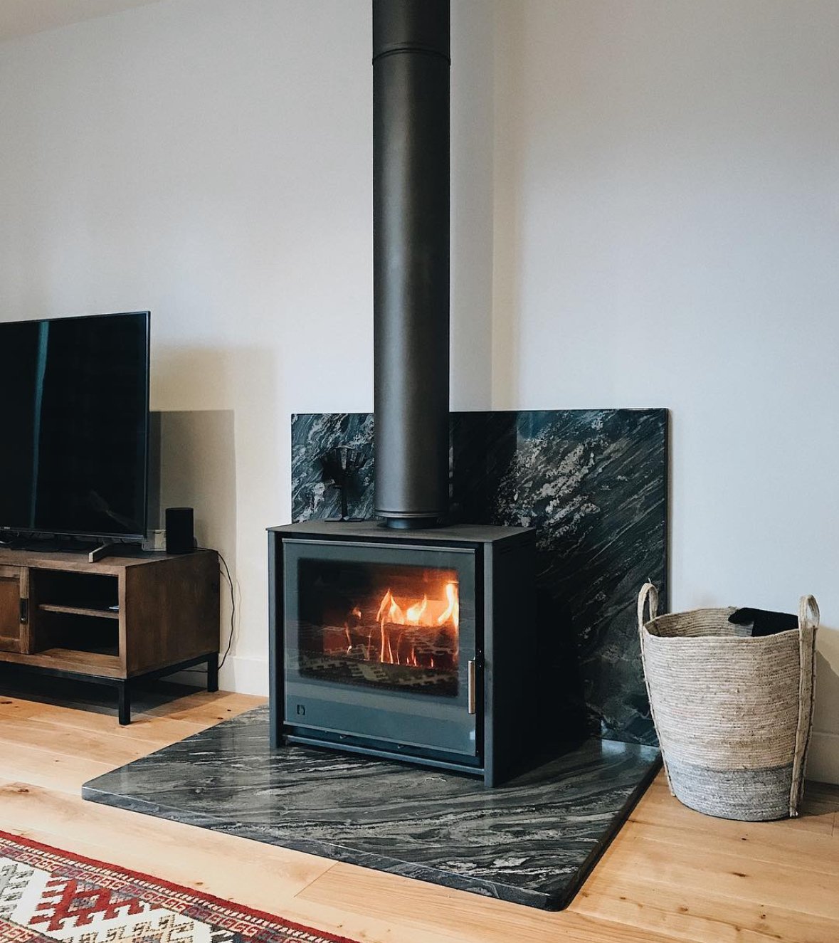 A modern living room featuring a black wood-burning stove with a visible fire inside, set on a dark marble hearth, with a basket of firewood next to it, a flat-screen TV on a wooden stand, and a colorful patterned rug partially visible in the foregro