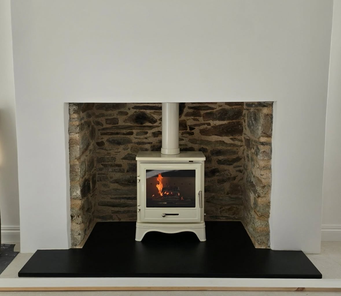 White wood stove with glass door in a stone fireplace with a black hearth and white mantel.