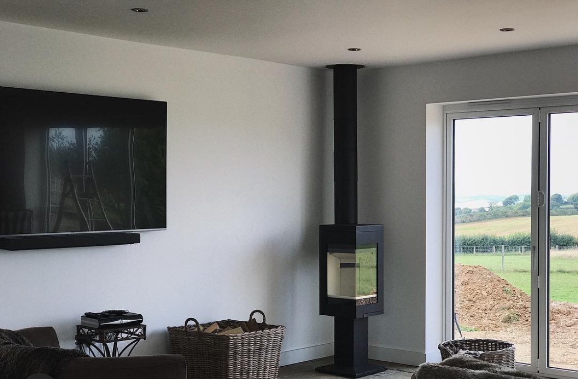 Living room with flat-screen TV on white wall, black fireplace, sliding glass door showing outdoor field, wicker laundry basket, small side table, couch, and pet bed.