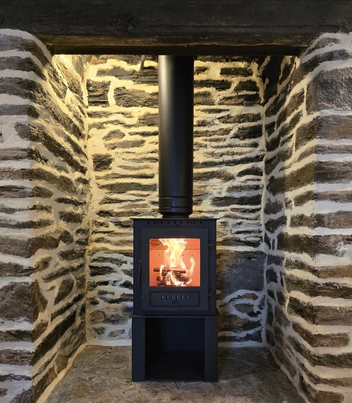 A black wood-burning stove with a visible fire inside, set against a wall of stacked stone and a stone floor.