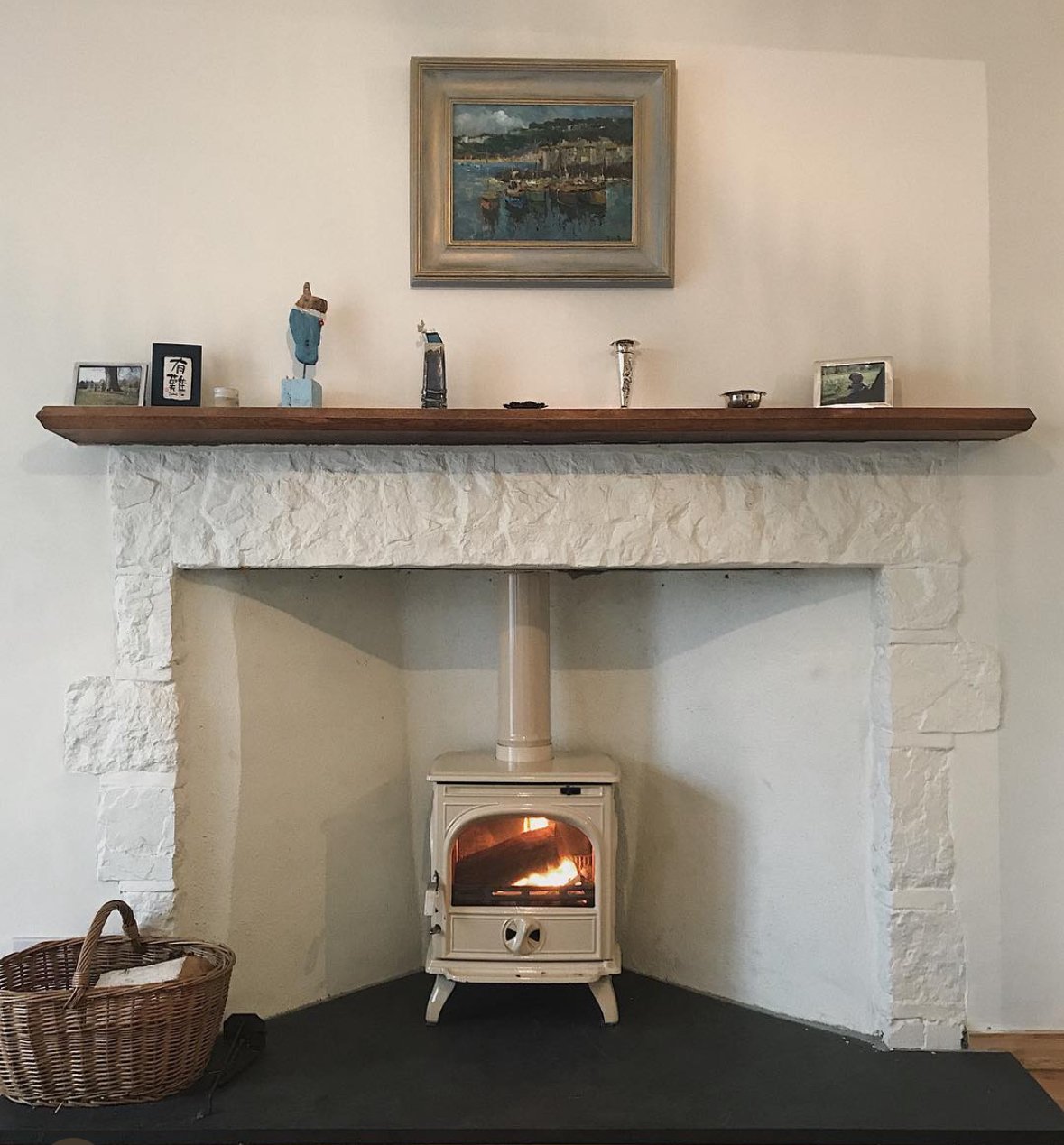 Living room fireplace with a wood stove burning, a basket of firewood on the floor, and a wooden mantel with small decorative items and framed photos; painting hanging above the mantel.