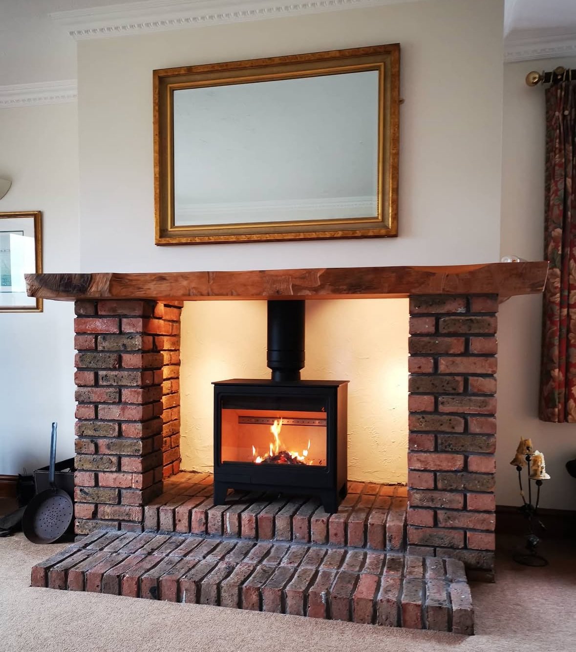 A brick fireplace with a wooden mantel, a wood stove with flames inside, and a large mirror with a gold frame hanging above it in a living room.