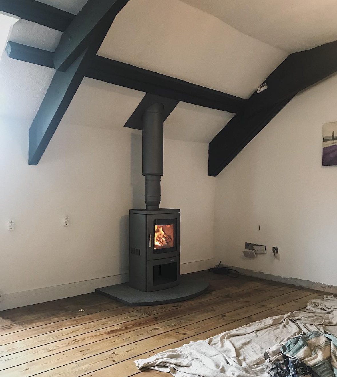 Interior of a room with a wood stove emitting a fire, positioned on a semi-circular hearth, with white walls, wooden floor, and black exposed beams on the ceiling.