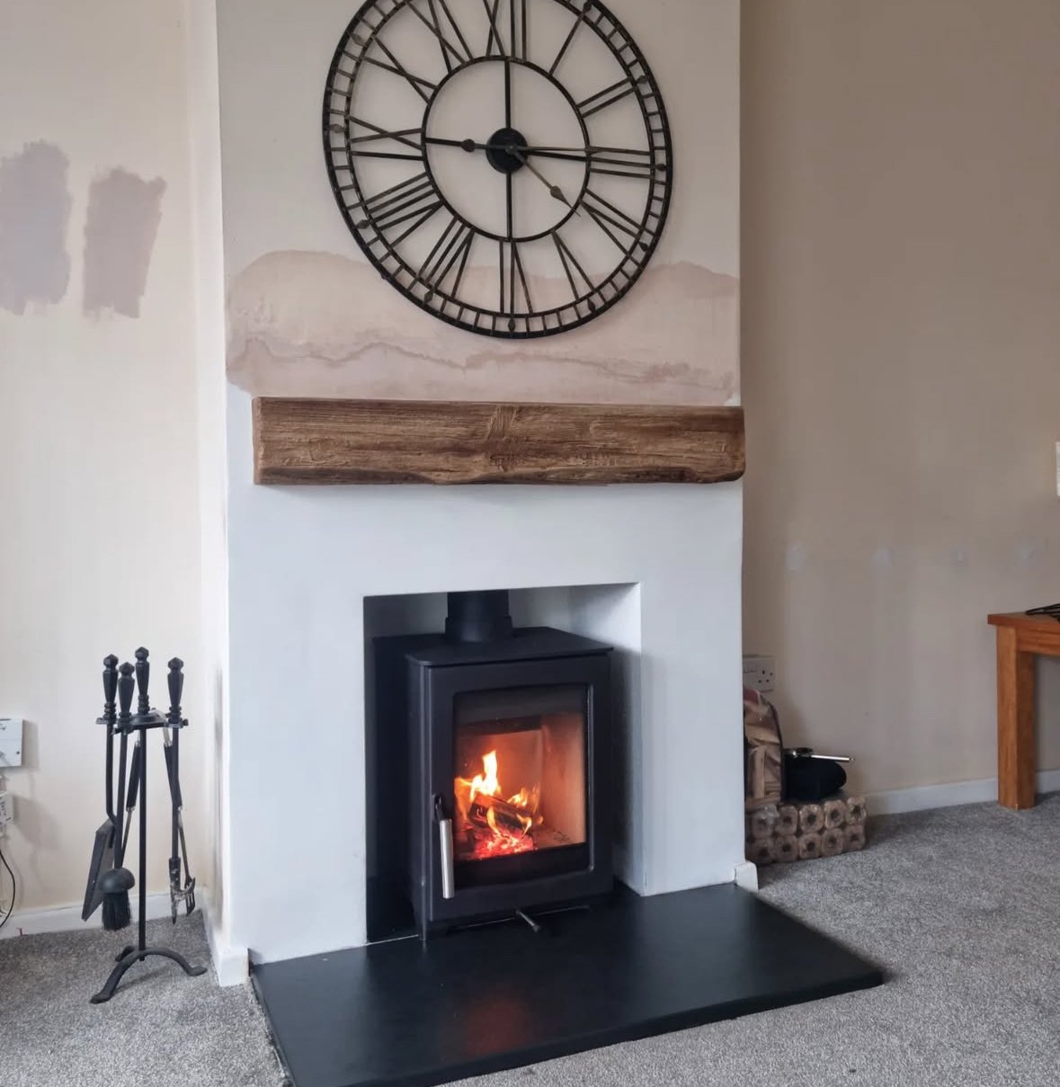 Living room fireplace with a wood stove, a large clock above, and a wooden mantel. To the left, fireplace tools, and to the right, a table and some rolled logs.