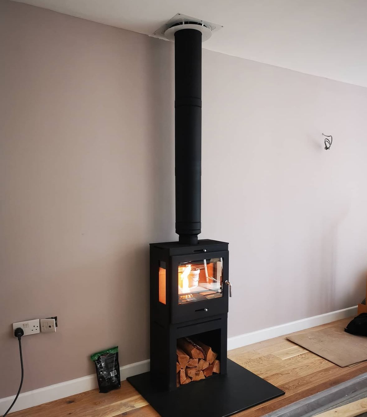 A black wood-burning stove with a glass door, connected to a vertical chimney pipe, situated on a black hearth in a room with light-colored walls and wooden flooring.