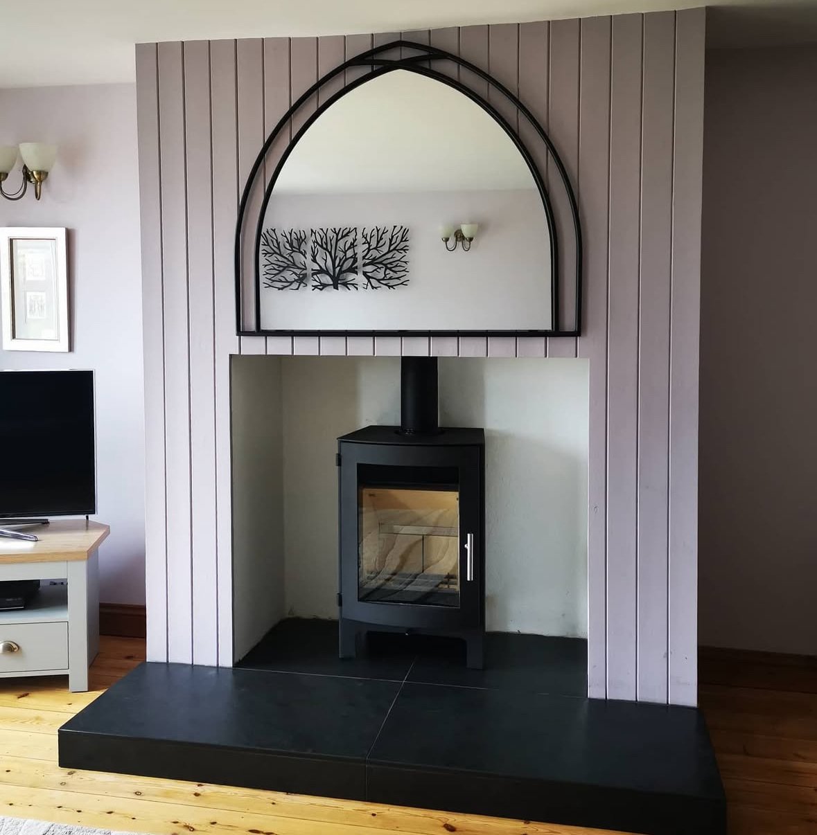 A living room fireplace area with a black wood stove, a large decorative mirror with a black arched frame above it, and light-colored vertical wood paneling on the wall.