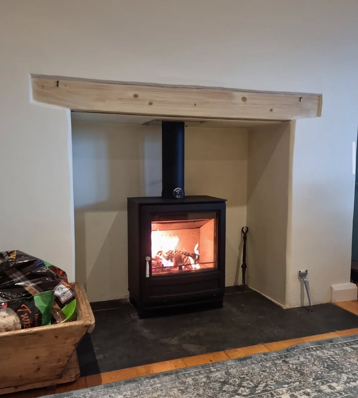 Wood stove with a fire burning inside, located within a recessed wall space with a wooden mantel above, a fireplace tool next to it, on a black hearth surrounded by a rug and wooden flooring.
