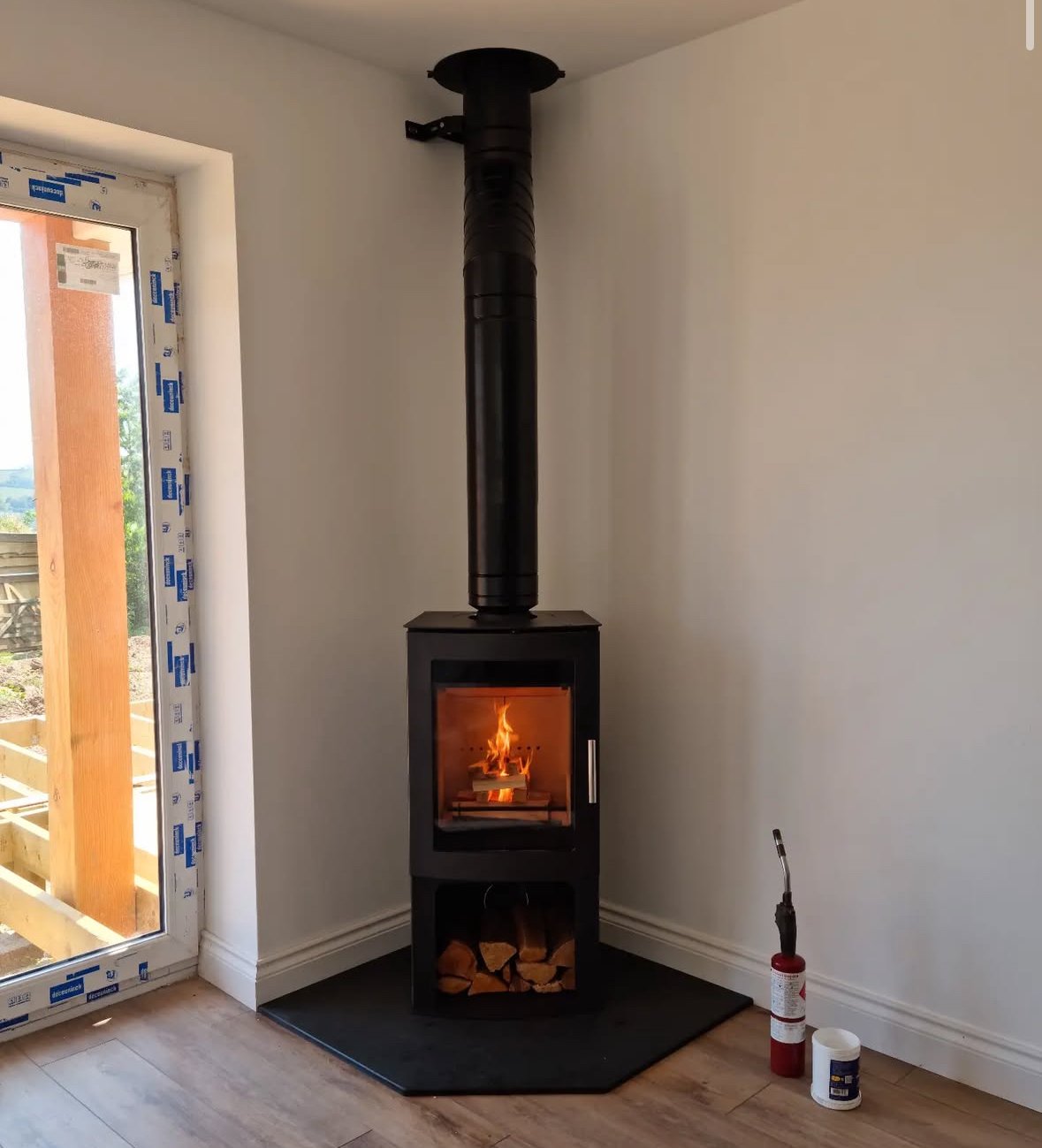 A black wood stove with a glass door showing a fire on a black mat on a wooden floor in a room with white walls, next to a sliding door with a view of a wooden deck outside.