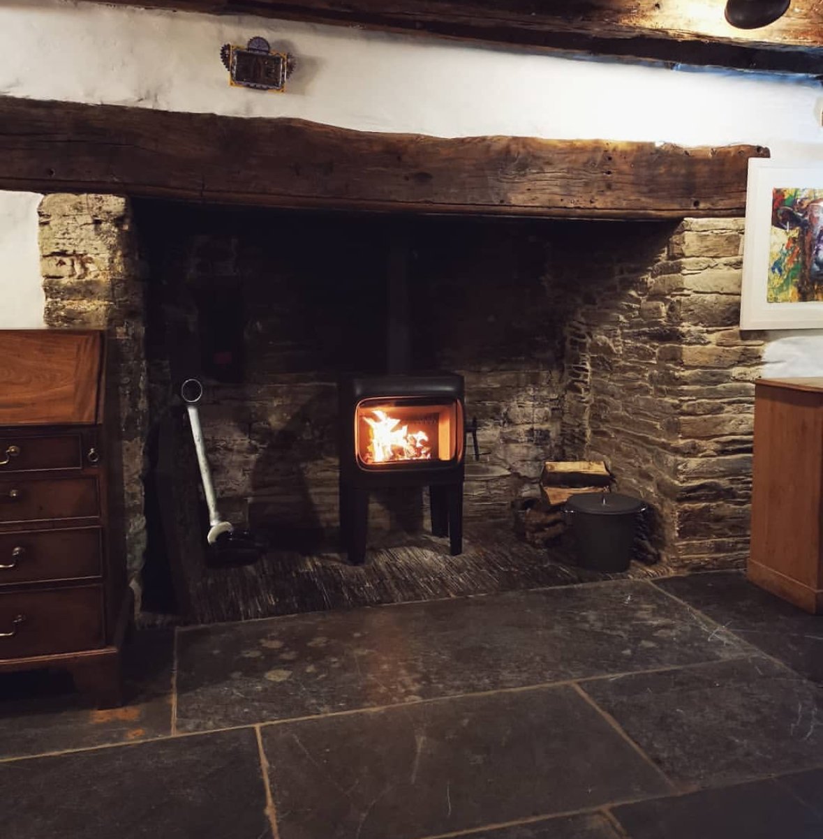 A cozy fireplace with a small burning fire inside, set in a rustic stone and wood mantel. There's a woodpile to the right, a metal bucket nearby, and antiques on the left, with a colorful painting on the right wall.