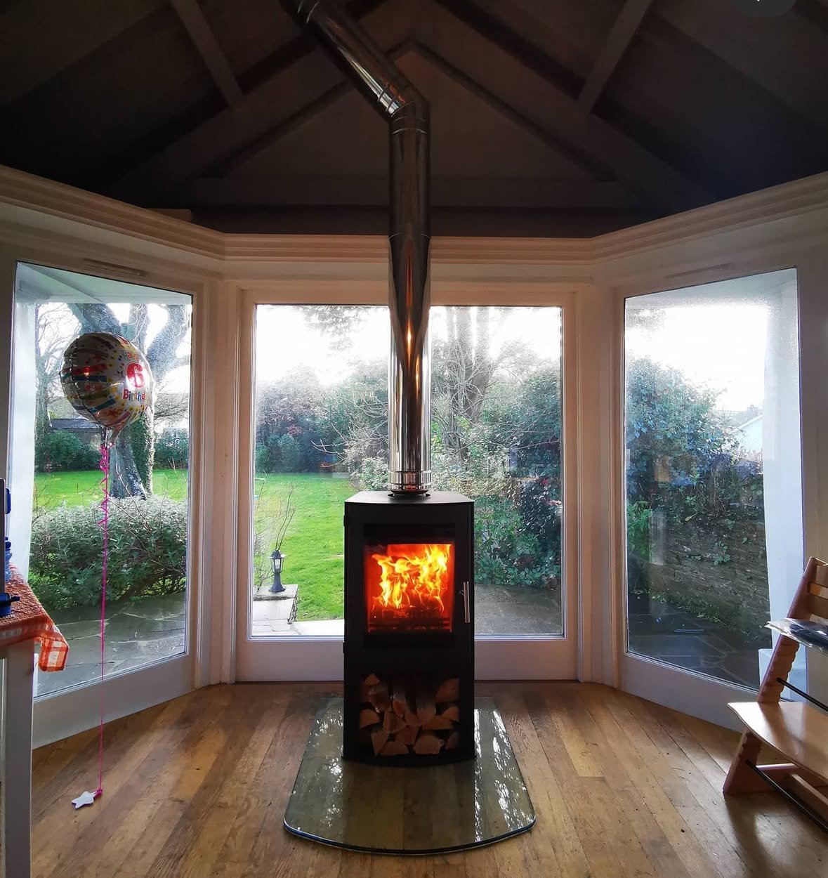 A wood-burning stove with a glass door showing flames, positioned in a sunroom with large bay windows overlooking a garden, with a small pile of firewood underneath, and a birthday balloon on the left side.