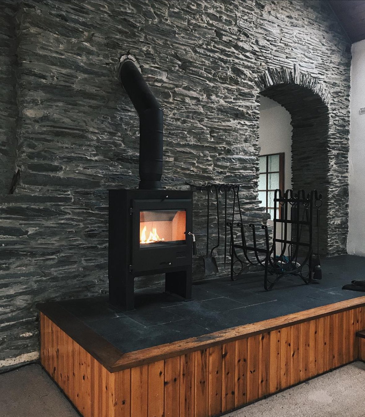 A cozy indoor fireplace with a black wood stove on a stone and wood platform, set against a textured stone wall with fireplace tools hanging nearby.