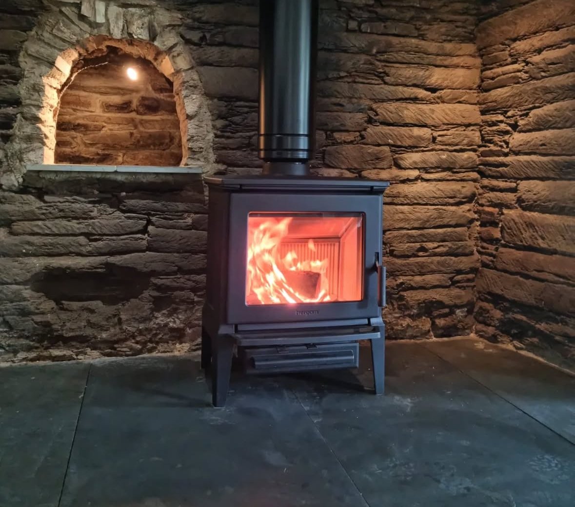 A black wood stove with a visible fire inside, positioned against a stone wall. There is a small arched niche in the wall above the stove.