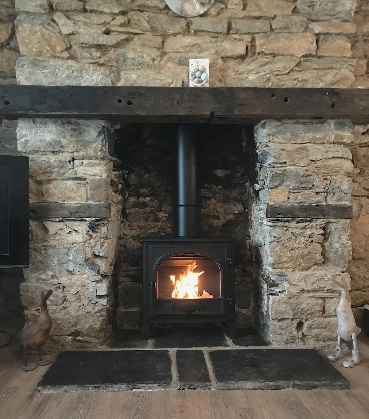 A stone fireplace with a lit wood stove inside, decorated with a small photo and figurines on the mantel. The fireplace has a rustic appearance with wooden and stone elements.