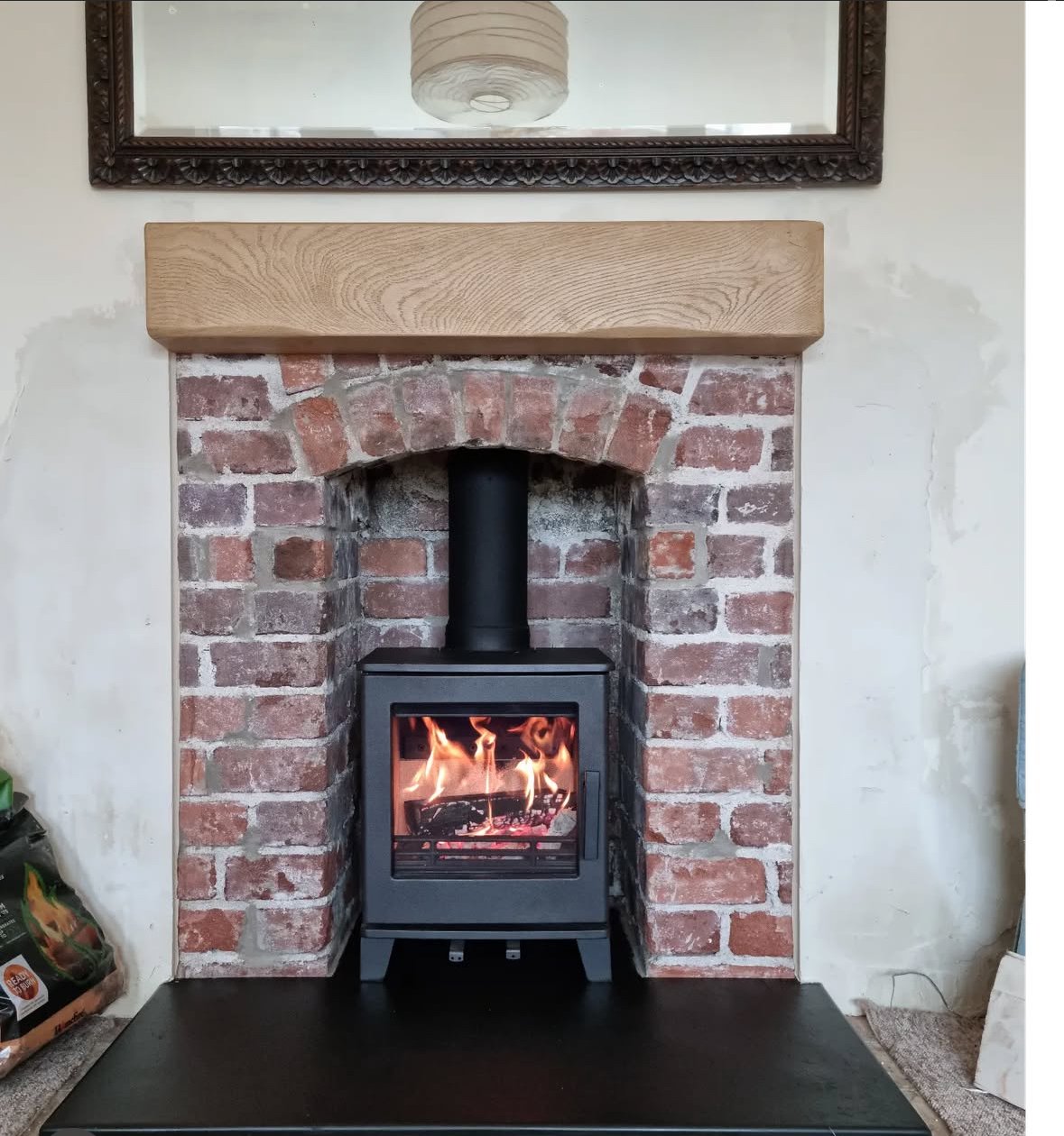 Wood-burning stove in a brick fireplace with a wooden mantel, with a framed mirror above and a paper lantern reflection visible.