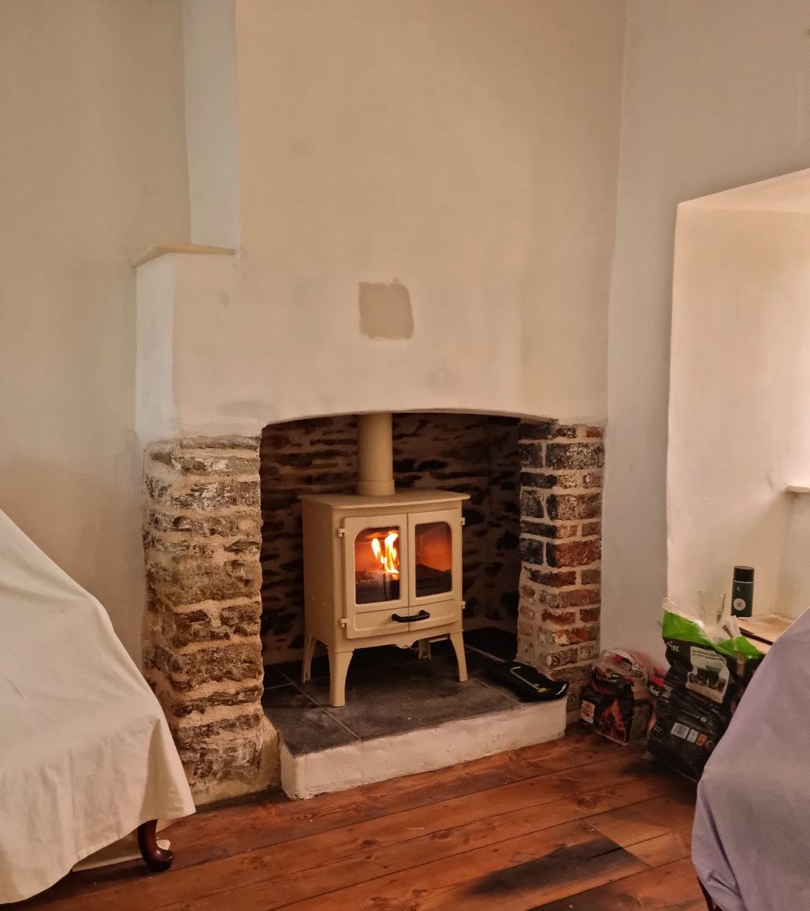 A cozy living room with a small cream-colored wood stove in a brick fireplace, burning fire inside. The room has wooden flooring and clutter on the right side, with part of a white draped piece of furniture on the left.