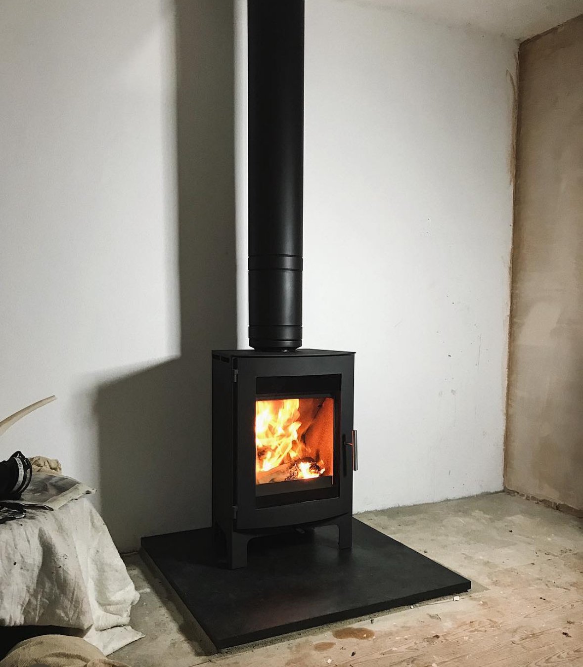 A black wood stove with a glass door showing a fire inside, connected to a black chimney pipe, installed on a black hearth pad in a room under construction with unfinished walls and flooring.