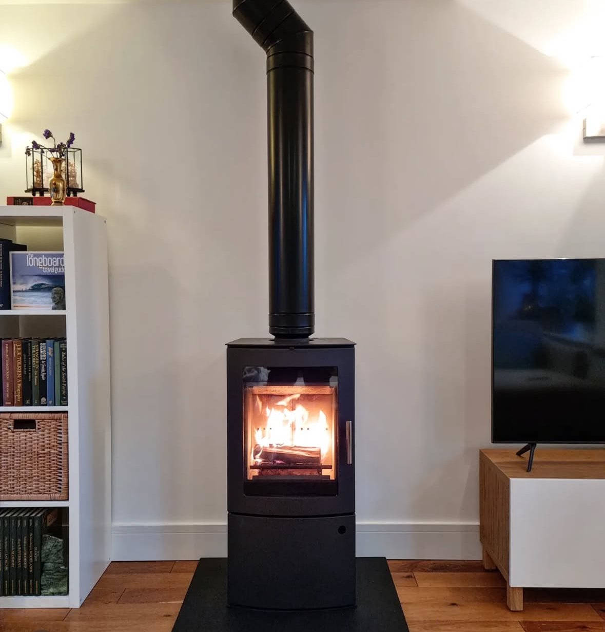 A black wood-burning stove with a visible fire inside, connected to a black chimney pipe extending up the plain white wall in a living room. To the left, a white bookshelf with books and decorative items, and to the right, a flat-screen TV on a woode