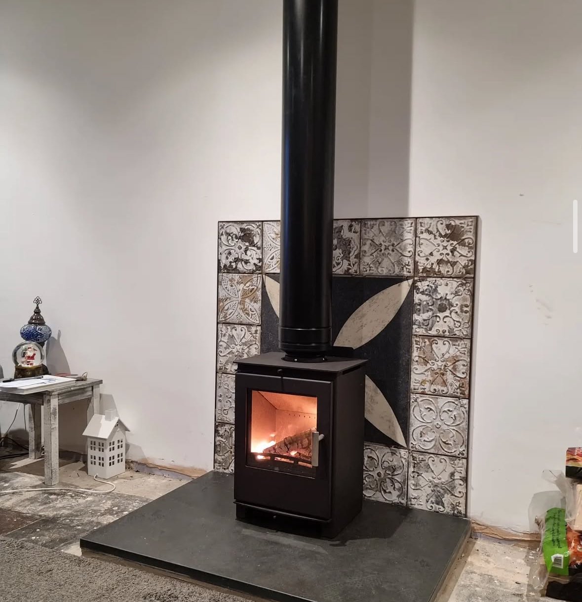 A black wood stove with a glass door showing a fire inside, positioned on a raised black platform, with decorative tile behind it and minimal furniture and decor nearby.