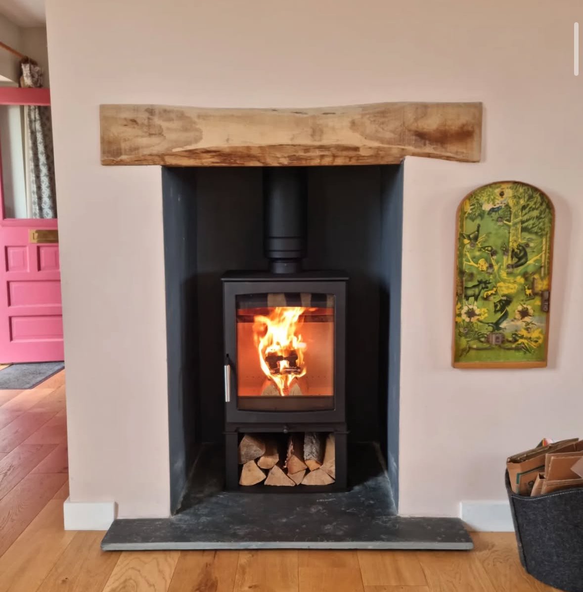 A wood-burning stove with a glass door showing a fire inside, set within a wall alcove with a wooden mantel above and a wood storage compartment below, positioned on a dark stone hearth.