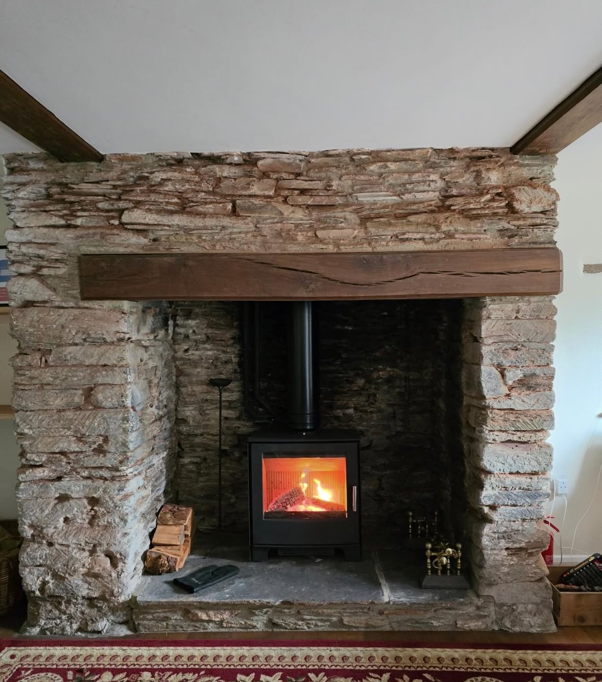 A cozy fireplace with a wood stove set inside a rustic brick surround and a thick wooden mantle above it.