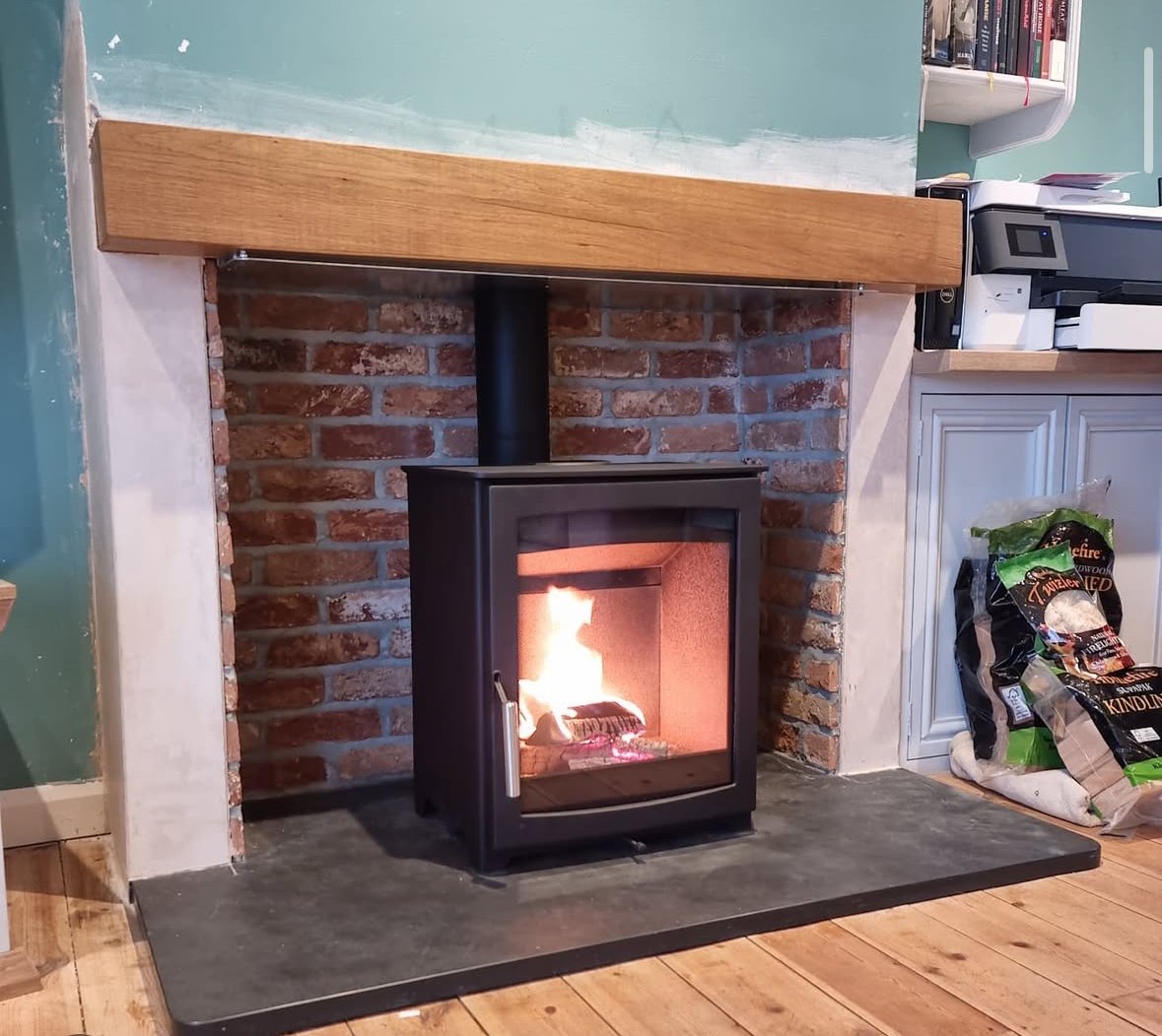 A black wood stove with a fire inside, positioned on a black hearth pad in front of a brick and painted wall. Part of a wooden mantel is visible above, and to the right, shelves, books, and a bag of wood pellets are seen.