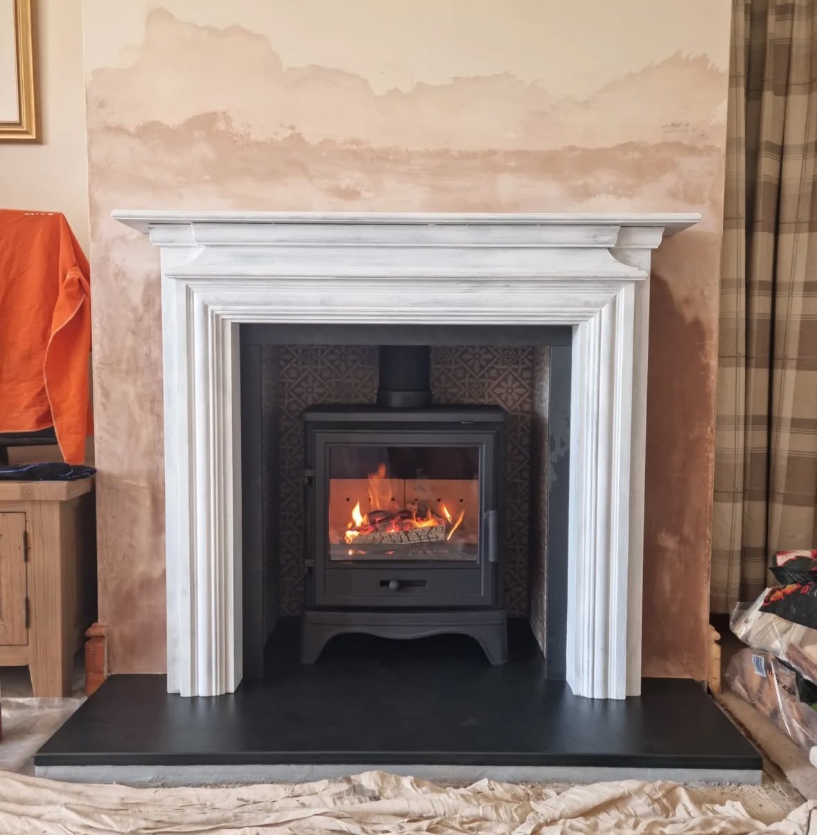 White fireplace mantel with a small black stove and fire inside, set against a partially painted wall and surrounded by construction or renovation materials.
