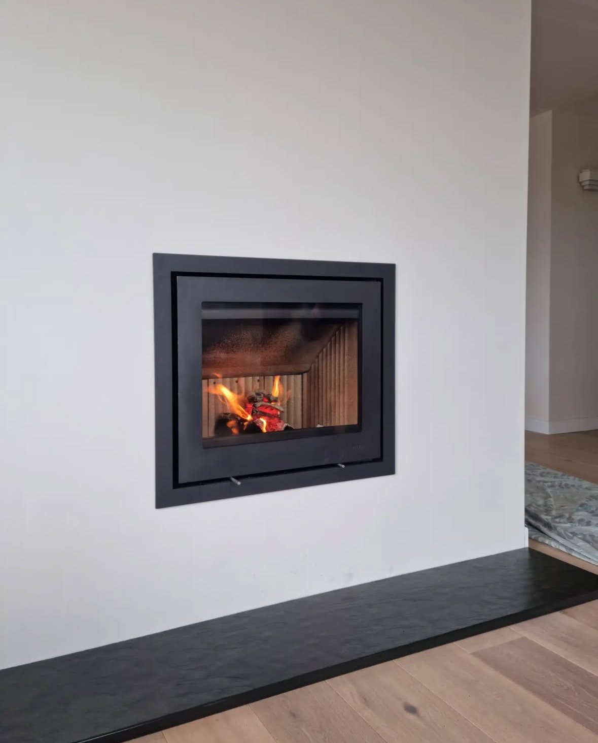 Modern living room corner with a built-in fireplace, black frame, white wall, wooden floor, small patterned rug, and a dark stone hearth.
